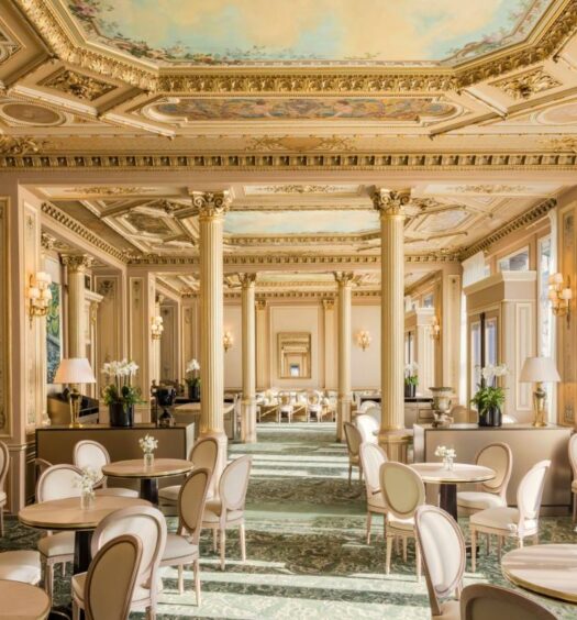 Salão de jantar do InterContinental Paris Le Grand, an IHG Hotel, um dos hotéis 5 estrelas em Paris. O local sofisticado tem pilastras e um teto abobadado com ilustrações renascentistas. Várias mesas e cadeiras estão distribuídas por ali.