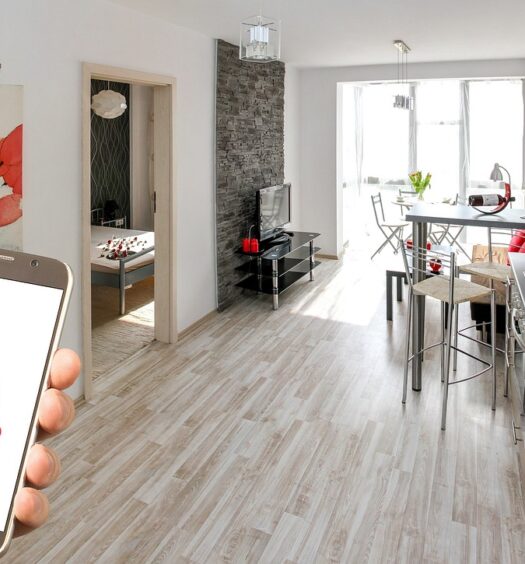 Mão segurando um celular com o logo do airbnb e, no fundo, é o interior de um apartamento. Há uma pia, balcão com bancos e a sala com TV atrás. Há, também, uma porta do quarto mostrando parte da cama