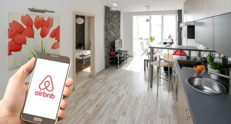 Mão segurando um celular com o logo do airbnb e, no fundo, é o interior de um apartamento. Há uma pia, balcão com bancos e a sala com TV atrás. Há, também, uma porta do quarto mostrando parte da cama