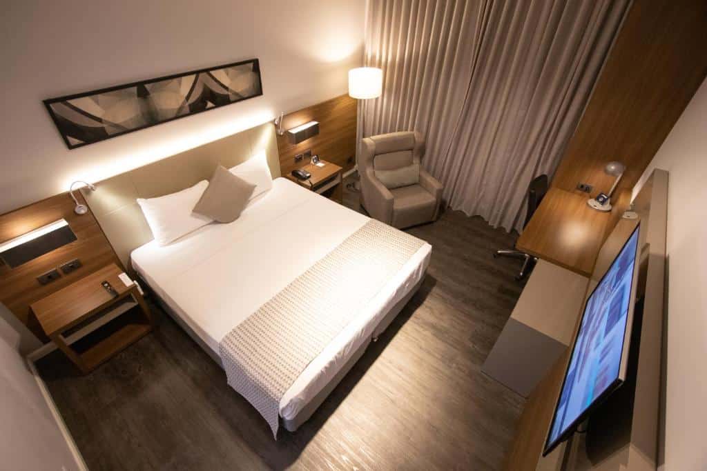 Foto do quarto do JL Hotel by Bourbon, ilustrando o post sobre Onde ficar em Foz do Iguaçu. A foto foi tirada de cima do quarto, mostrando embaixo uma cama box de casal, uma poltrona na direita, e uma TV e escrivaninha na frente da cama..
