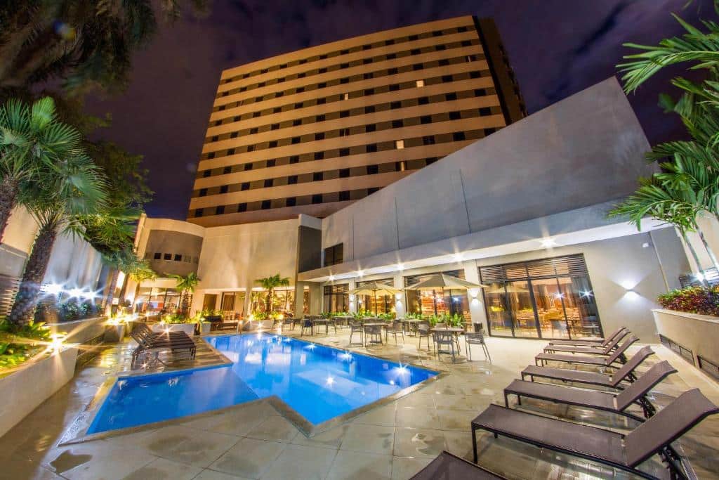 Foto do JL Hotel by Bourbon, mostrando um deck com piscina, espreguiçadeiras, mesas com guarda-sol. No fundo se vê o hotel, e está de noite.
