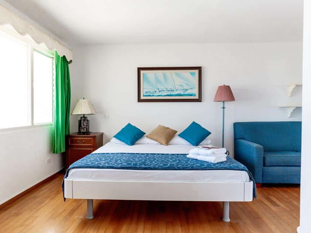 Quarto do Kamerlingh Villa. Do lado esquerdo uma cômoda com abajur e uma janela com cortina. No meio uma cama de casal. Do lado direito um sofá e um abajur. Foto para ilustrar post de onde ficar em Aruba.