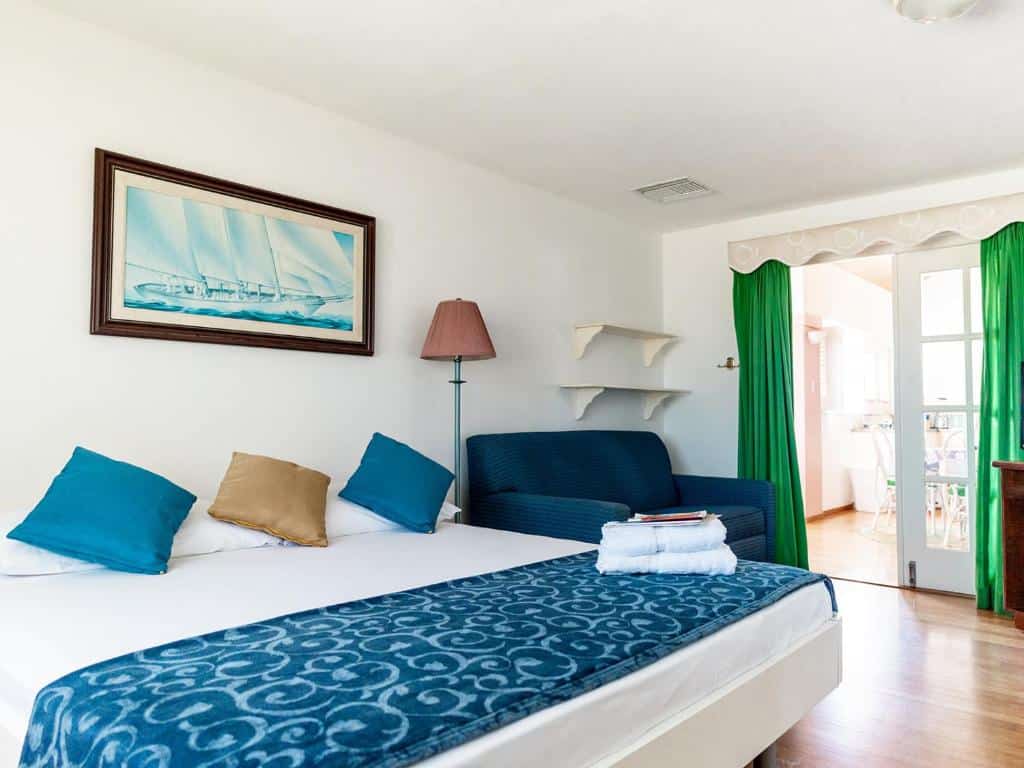 Quarto do hotel Kamerlingh Villa. Uma cama de casal está do lado esquerdo da imagem. Ao seu lado direito e embaixo de duas prateleiras na parede há uma poltrona grande. Depois da poltrona está uma porta para outro cômodo. Este é um dos hotéis baratos em Aruba.