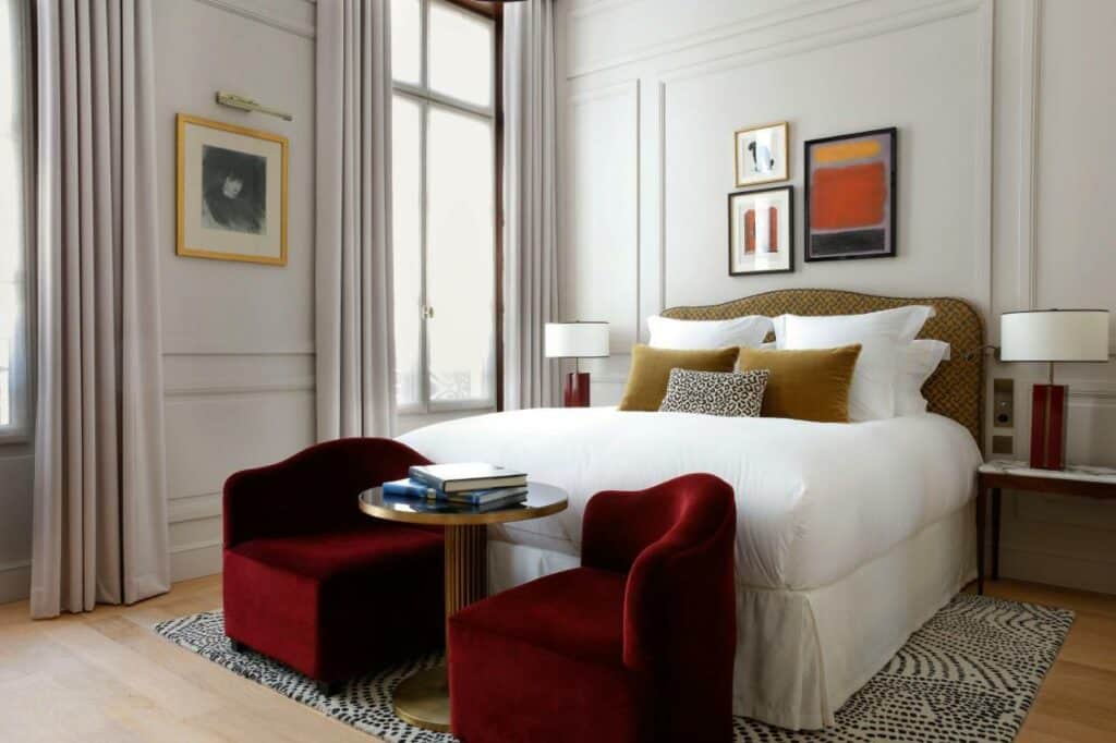 Quarto do Les Jardins du Faubourg Hotel & Spa by Shiseido, um dos hotéis 5 estrelas em Paris. Uma cama de casal está com mesinhas de cabeceira e abajures dos dois lados está encostada na parede central, e aos seus pés ficam uma mesinha e duas poltronas. Há um tapete embaixo da cama. Na parede ao lado esquerdo da cama ficam duas janelas com cortinas.