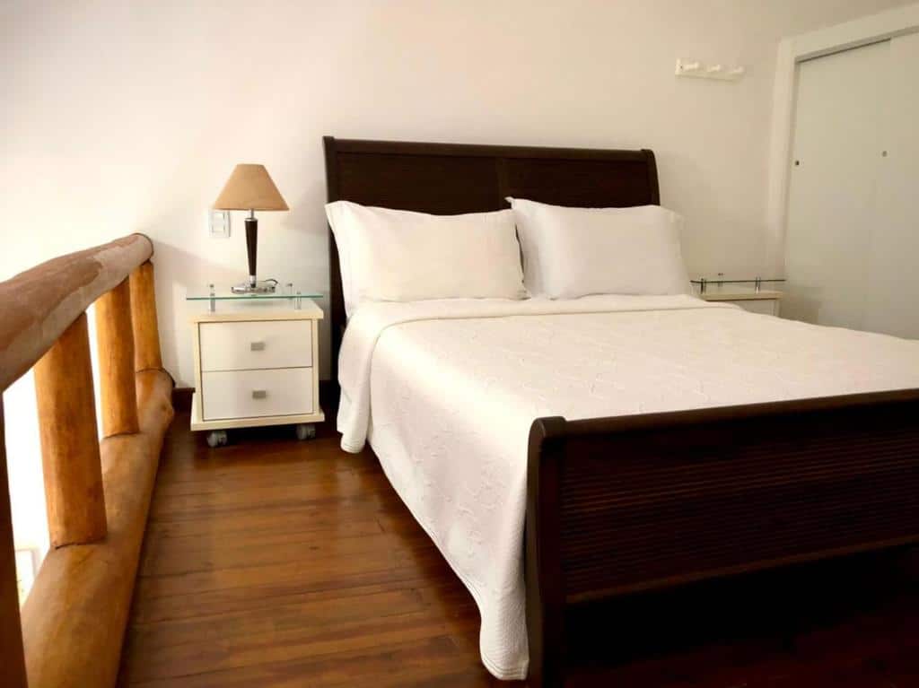 Foto do quarto em Loft encantador em Praia do Forte próximo à Vila, que ilustra o post de airbnb na Praia do Forte. Há uma cama de casal em cima do segundo andar do loft, que possui barras de madeira. Dos dois lados da cama há uma mesa de cabeceira branca com gavetas, e a da esquerda possui um abajur em cima.