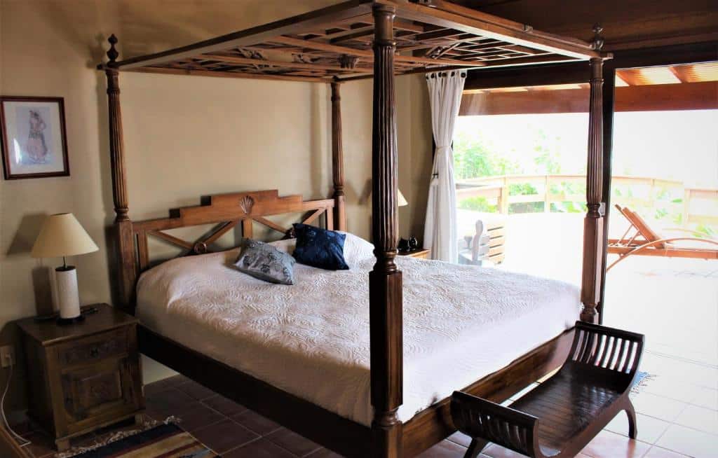 Quarto da Luxury Suites Vila Indonésia, airbnb em Garopaba. Uma cama de casal com dossel está encostada na parede ao lado esquerdo e tem mesinhas de cabeceira com abajures dos dois lados, assim como um banco de madeira aos pés. Na lateral direita fica uma ampla porta de vidro que leva à varanda, onde é possível ver uma espreguiçadeira.