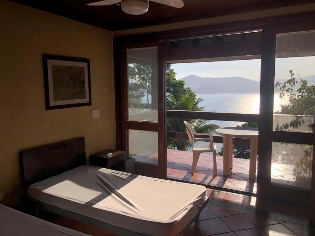 Quarto da Mansão na costa com vista para o mar. Do lado esquerdo duas camas de solteiro, no fundo a porta da varanda com mesa e cadeira e vista do mar. Foto para ilustrar post sobre airbnb na Praia da Enseada.