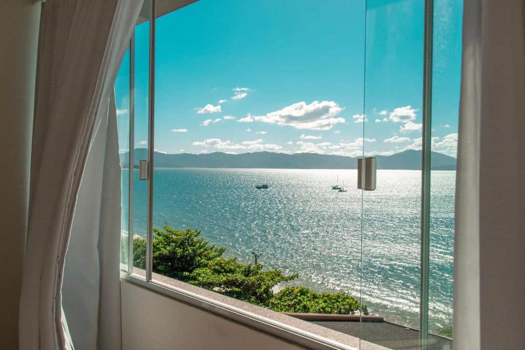 Foto tirada da janela do airbnb Mergulho no Mar de Jurerê com Vista Frontal. A janela oferece vista para o mar, com faixas de terra ao fundo e o céu. Pode-se ver também algumas árvores.