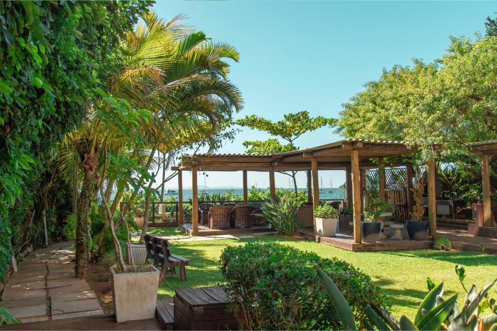 Área externa do airbnb Mergulho no Mar de Jurerê com Vista Lateral. Há uma grande área verde, cercada por árvores e arbustos. Ao fundo é possível ver uma estrutura com vigas e um teto de madeira. Nessa estrutura há cadeiras, mesas e algumas plantas em vasos. No horizonte pode-se ver o mar.