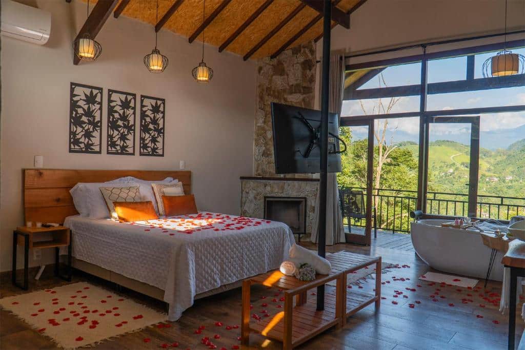 Imagem do quarto em Monte Mauá Chalés, que está ilustrando o post sobre airbnb em Visconde de Mauá. Há uma cama box de casal no centro do quarto, com pétalas de rosa espalhada por cima dela e do chão do quarto. Do lado direito da cama há uma lareira, varanda e também uma banheira. O ambiente é espaçoso. Uma TV está fixa em uma coluna de frente para a cama.