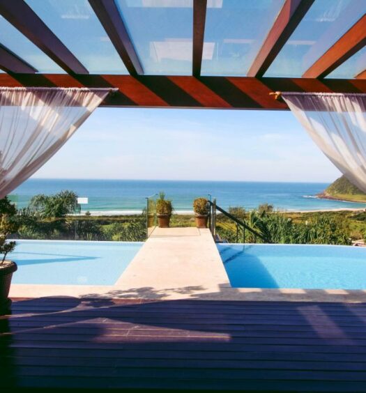 Visão da piscina da Morada Prainha, um dos airbnb em Garopaba. Uma cobertura de madeira com cortinas brancas leva ao deque da piscina, que tem vista direta para a praia em frente.