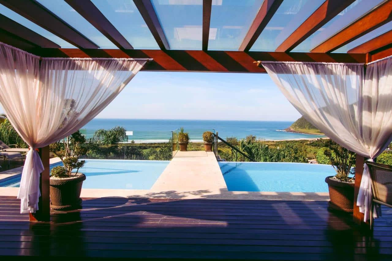 Visão da piscina da Morada Prainha, um dos airbnb em Garopaba. Uma cobertura de madeira com cortinas brancas leva ao deque da piscina, que tem vista direta para a praia em frente.