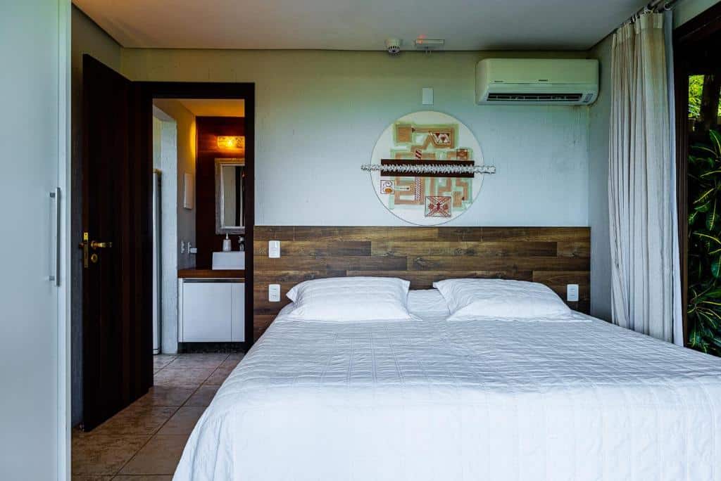 Quarto da Morada Prainha, airbnb em Garopaba. Uma cama de casal encostada na parede central tem uma decoração de parede logo acima, assim como um aparelho de ar-condicionado. Ao lado esquerdo fica uma porta para o banheiro, e ao direito uma janela com cortinas.