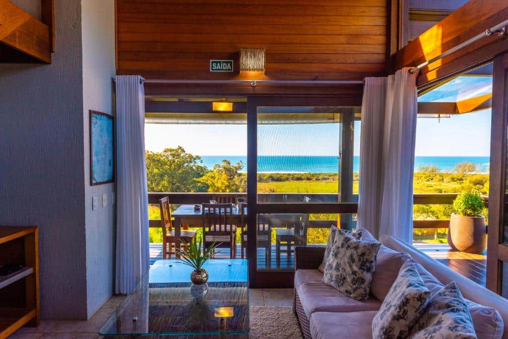 Sala da Morada Prainha. Um sofá ao lado direito da imagem encara uma mesinha de centro e um suporte com TV na parede oposta. As paredes do local são tomadas pelas portas de vidro que levam à varanda com mesa com cadeiras e vista para a praia.