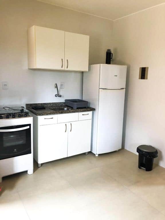 Cozinha das Moradas Costta Sul. No centro da parede há uma bancada com pia e um armário suspenso logo acima. Ao lado esquerdo fica o fogão e ao direito, a geladeira com uma garrafa de café em cima. Em frente à geladeira há uma lata de lixo.