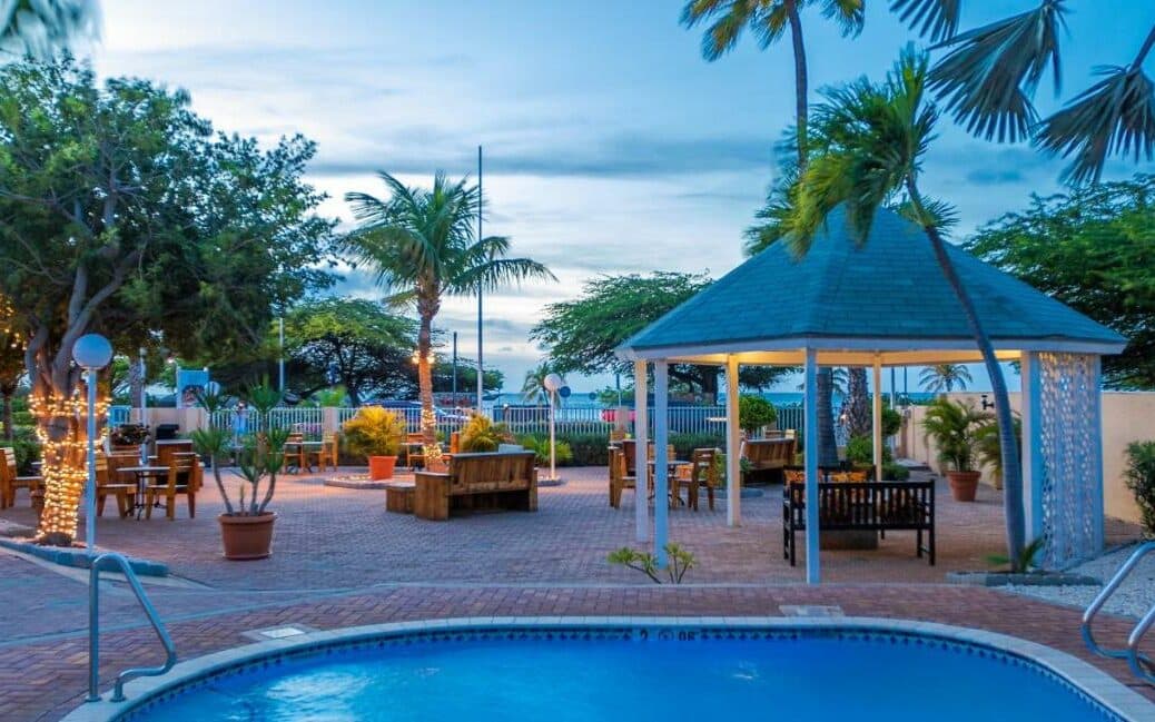 Área externa do hotel MVC Eagle Beach. Uma piscina está no canto da imagem. Na frente dela há um gazebo e diversos bancos espalhados pelo ambiente. Algumas árvores também decoram o local.