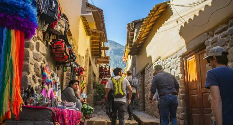 homem andando pelas ruas de cusco. Ele está bem no centro da imagem, de costas para a câmera, olhando para a direita. Em cada lado da rua há construções, sendo que no lado esquerdo, na parte inferior, há uma mesa repleta de artesanatos diversos, ao lado de uma mulher