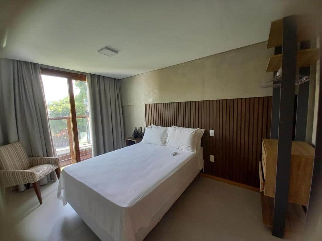 Foto do quarto em Ohana Praia do Forte, que ilustra o post de airbnb na Praia do Forte. O quarto é grande e moderno. No centro há uma cama de casal, na direita da cama há uma estante com gavetas, e na esquerda há uma porta de vidro com varanda e uma poltrona no canto.