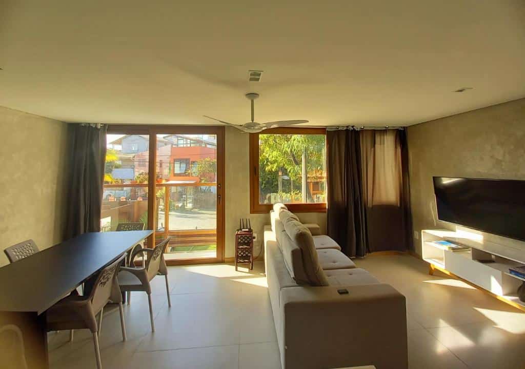 Foto de uma sala na Ohana Praia do Forte. Mais pra a direita está o sofá e uma TV grande à sua frente, e atrás do sofá, já na esquerda, há uma mesa extensa com cadeiras para jantar. Essa sala é bem iluminada com uma janela ao lado do sofá e uma porta de vidro do lado da janela.