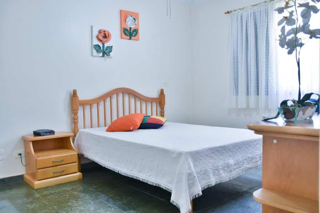 Imagem do quarto do Orange Beach Enseada Guarujá, ilustrando o post sobre airbnb em Guarujá. Há uma cama de casal no centro com almofadas em cima, e do lado esquerdo dela há uma mesa de cabeceira com gavetas. No canto inferior direito da foto há uma cômoda com vaso de plantas de frente para a cama. E do lado direito há uma janela.