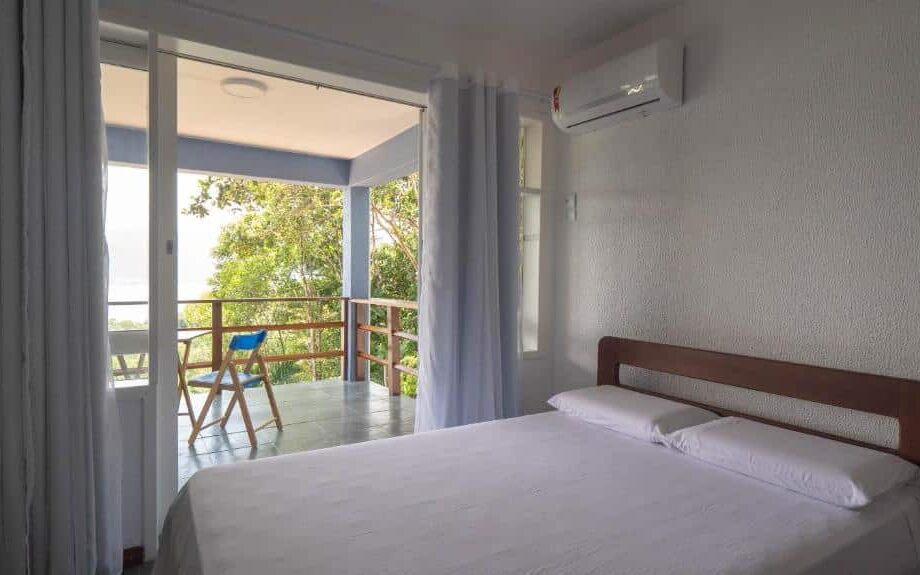 Quarto do Oré Suítes e Chalés. Ao lado direito está uma cama de casal. Do lado esquerdo está a varanda com uma mesa e cadeiras e rodeada de árvores. Este é um dos airbnb na Praia Vermelha do Centro.