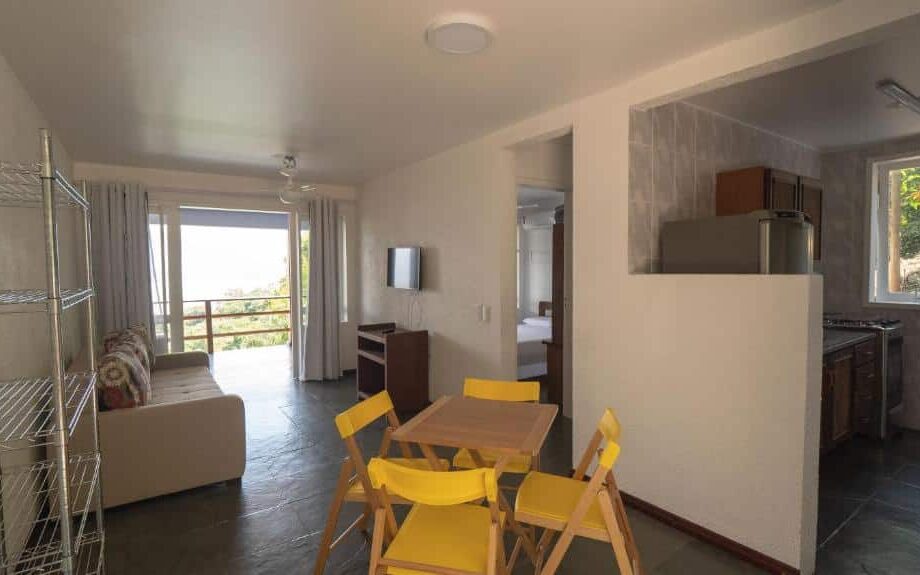 Sala e cozinha da Praia Vermelha do Centro. A sala está ao lado esquerdo, com um sofá de frente para uma tv e a varanda ao fundo. Do lado direito está a cozinha, com uma pequena parede fazendo a divisória do espaço. Entre os dois cômodos há uma mesa de quatro lugares.