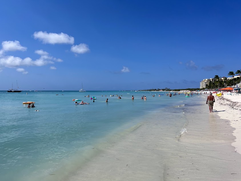 Um mar cristalino em Palm Beach do lado esquerdo, com várias pessoas no mar. Algumas pessoas caminhando na beira do mar e a areia do lado direito.