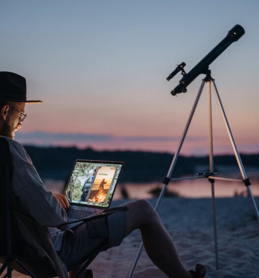 Um rapaz sentado em um local com areia, há um telescópio, e ele está sentado em uma cadeira com um notebook no colo, para representar seguro viagem nômade digital