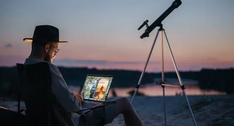 Um rapaz sentado em um local com areia, há um telescópio, e ele está sentado em uma cadeira com um notebook no colo, para representar seguro viagem nômade digital