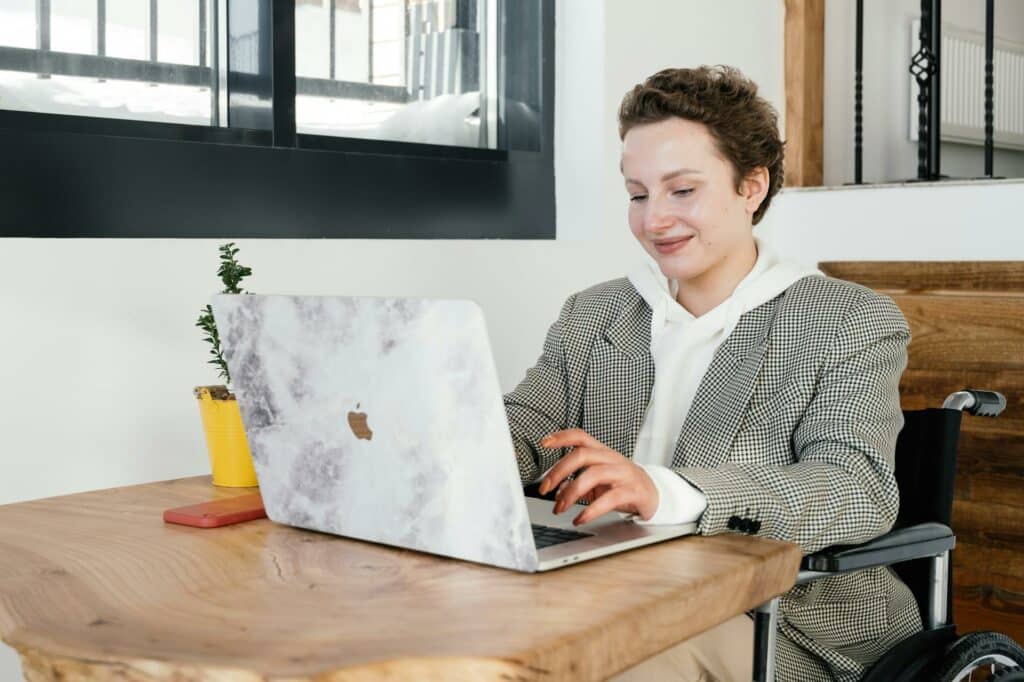 Uma mulher em uma cadeira de rodas próxima de uma mesa de madeira onda há um notebook aberto que ela está usando