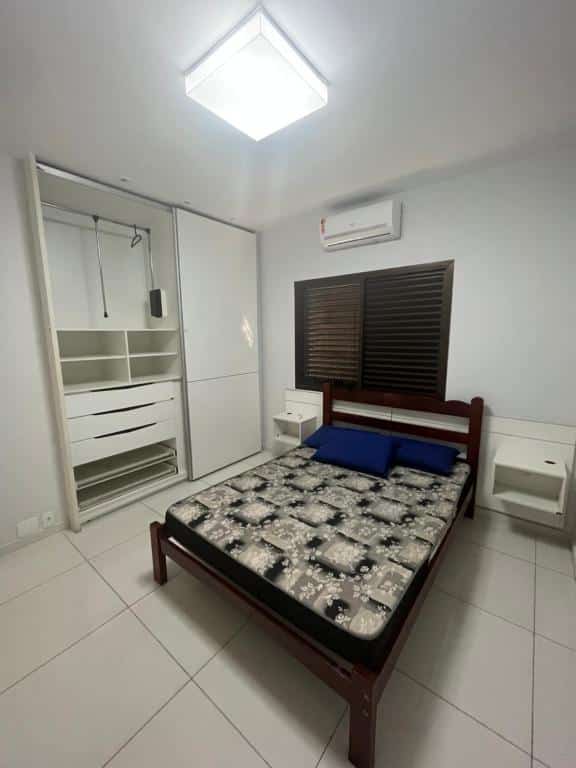 Imagem do quarto do  Pitangueiras Service, ilustrando o post sobre airbnb em Guarujá. No centro há uma cama de casal e uma janela e ar-condicionado atrás, e do lado esquerdo da cama há um guarda-roupa.