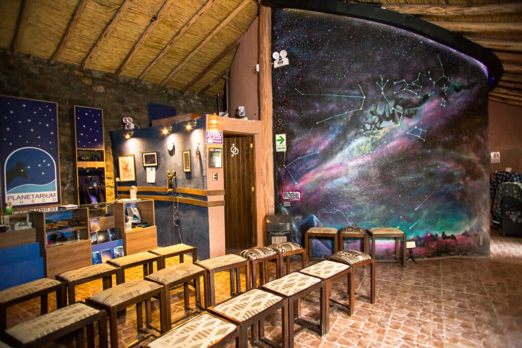 sala do planet[ario de Cusco com várias banquetas de madeira enfileiradas na parte inferior esquerda da imagem. No lado direito há uma grande parede abaulada, pintada com desenhos cósmicos. E ao fundo, atrás das banquetas, há uma galeria de souvenires.