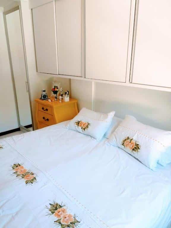 Foto do quarto em Porto Praia do Forte 2s, que ilustra o post de airbnb na Praia do Forte. As paredes, armários e roupa de cama são brancas. A cama é box e de casal, há armários em cima dela e ao lado há uma mesa de cabeceira com gavetas.