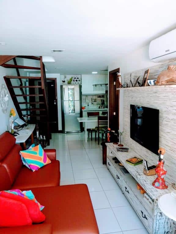 Sala na Porto Praia do Forte 2s. Há um sofá vermelho à esquerda, e na frente há um painel com televisão e um ar-condicionado no topo. No fundo há uma escada na esquerda e uma cozinha completa na direita.