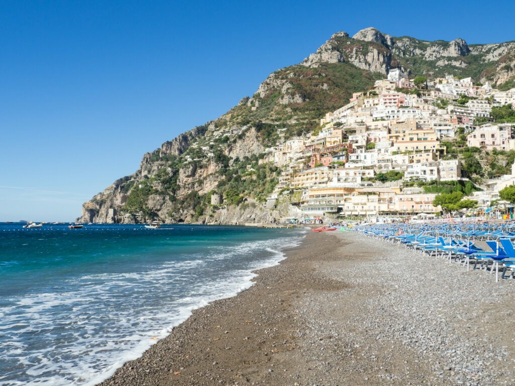 vista com uma faixa de areia dividindo a orla de um lado com cadeiras e águas claras do outro lado, ao fundo as construções em pedra em uma grande montanha na Costa Amalfi, em Positano, na Itália, que pode ser visitado com o CDAM