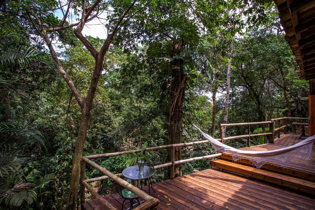 Varanda do quarto da Pousada Barulho D' água. Nesta imagem pode-se ver uma grande varanda com o piso feito em madeira e com parapeito de madeira. Na ponta esquerda da varanda há uma pequena mesa de vidro com vaso de planta em cima e duas cadeiras pequenas, ao centro da varanda é possível ver uma rede pendurada e logo a frente uma elevação com dois degraus. A varanda possui vista para uma vasta floresta.