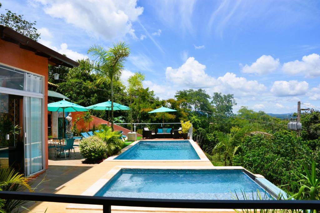 Área externa da Pousada Boutique Mirante d'Italia. Duas piscinas de bordas infinitas no meio, do lado direito árvores, do lado esquerdo cadeiras de tomar sol e guarda-sóis.