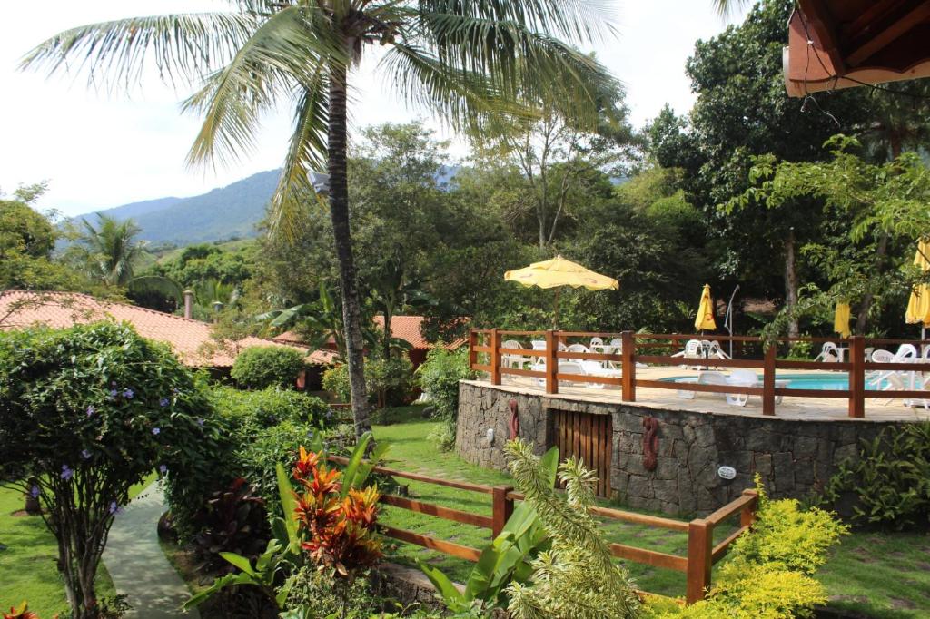 Área externa da Pousada Cantinho D'Abrantes. A direita é possível ver a estrutura da piscina, que é cercada por madeira. Em volta da piscina há muita grama, natureza, árvores e arbustos. A esquerda há um caminho de pedra entre alguns pequenos arbustos.