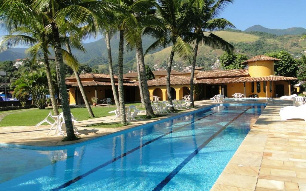 Área externa da Pousada Casa Amarela. Há uma grande piscina, cercada por espreguiçadeiras, cadeiras e árvores, ao fundo é possível ver uma grande casa amarela.