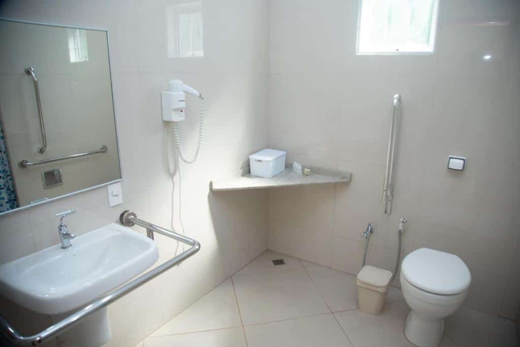 Banheiro da Pousada Casa Rosa com acessibilidade. Do lado direito um vaso sanitário com barras de apoio, no meio uma bancada, do lado esquerdo uma pia com barras de apoio, um espelho e um telefone.