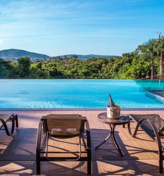Área externa da Pousada Cavaleiro dos Pireneus (Pirenópolis) para ilustrar post sobre pousadas em Goiás. Cadeiras de tomar sol na frente, no meio piscina de borda infinita com vista para a mata.