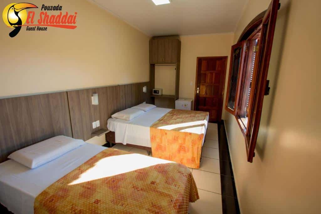 Foto do quarto da Pousada El Shaddai, ilustrando o post sobre Onde ficar em Foz do Iguaçu. Há duas camas de casal, encostada na parede à esquerda. Na frente delas há uma janela. No fundo do quarto há um armário, frigobar e porta.