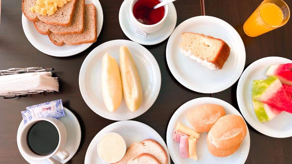 Foto da Pousada El Shaddai, mostrando de cima vários pratos com peças, frutas, frios, bolo, um copo de suco e uma xícara de café.