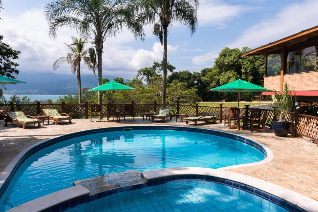 Área externa da Pousada Ilhote da Prainha. Pode-se ver ao centro duas piscinas, sendo uma maior e mais funda e a outra menor e mais rasa. Em volta da piscina há guarda-sóis, cadeiras, mesas e espreguiçadeiras, além de muitas árvores e natureza. Ao fundo é possível ver o mar.