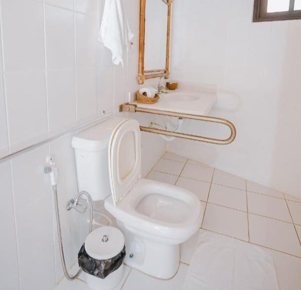 Banheiro acessível da Pousada Maná. Uma privada está ao lado de uma pia e espelho na parede. Entre eles há uma barra de apoio.