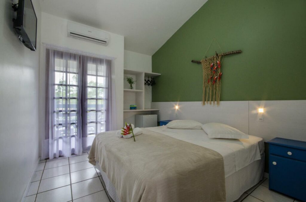 Quarto da Pousada Poente do Sol. Uma cama de casal está encostada na parede ao lado direito e conta com mesinhas de cabeceira e luminárias dos dois lados. Há uma decoração na parede de cabeceira, e uma TV na parede em frente. Na lateral esquerda da cama fica uma porta de vidro com cortinas que leva à varanda, e logo acima está um aparelho de ar-condicionado. Ao lado da porta há um armário com cabides e frigobar.