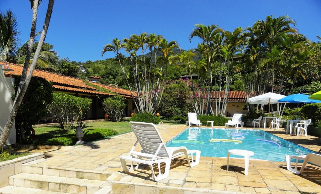 Área externa da Pousada Praia do Curral. Na direita está uma piscina cercada por cadeiras, mesas, espreguiçadeiras e guarda-sóis. A esquerda há uma faixa de grama com plantas, arbustos e uma casa. Ao fundo há muitos coqueiros, árvores e plantas.