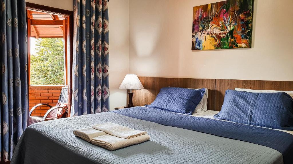 Foto a Pousada Rio Das Águas, mostrando uma cama de casal na direita, ocupando o centro da foto. Atrás da cama há uma parede com um quadro abstrato. Na esquerda da cama há um abajur e uma porta que dá passada para uma varanda com vista.