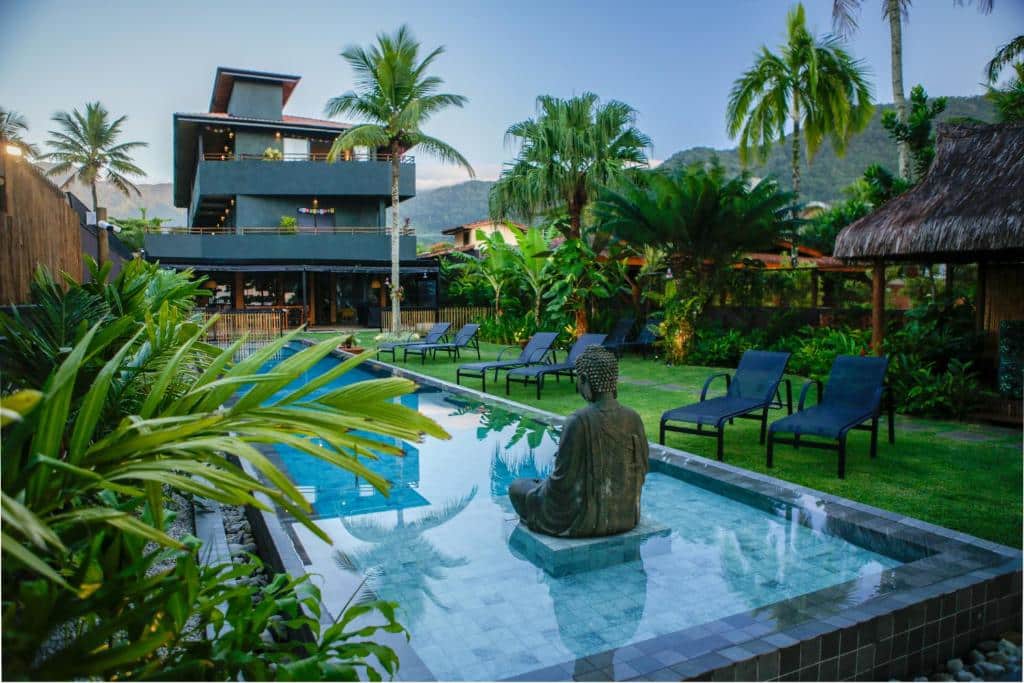 Parte da Pousada TeMoana que mostra a piscina ao ar livre com uma estátua na parte rasa da piscina. Em volta tem algumas plantas, espreguiçadeiras e um amplo gramado verde. Já ao fundo tem a construção do resort com três andares. Imagem ilustrando post Airbnb em Picinguaba.