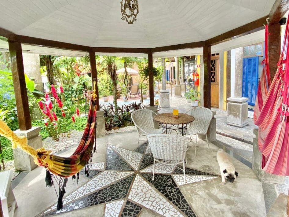 Área externa da Vila Pequeá. Na frente da imagem há uma estrutura circular coberta, no lado esquerdo e no lado direito há redes. No meio há uma mesa circular com três cadeiras de palha e um cachorrinho passando ao lado. No fundo, ao lado esquerdo há um jardim e no lado direito há portas coloridas.